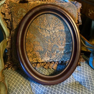 May include: A dark brown, oval picture frame. The frame is empty, revealing a patterned fabric in shades of teal, gold, and brown. The fabric is part of a decorative pillow with gold tassels. The frame is resting on a patterned cushion.