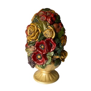 Könnte beinhalten: Keramik-Blumenarrangement-Skulptur in einer Sockelvase. Die Skulptur zeigt eine Vielzahl von Blumen in Rot-, Gelb- und Grüntönen. Die Vase und der Sockel sind hellbeige. Zu den Blumen gehören Rosen und Mohnblumen.