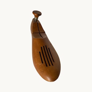 Puede incluir: Una horma de zapato de madera de color marrón claro con acabado liso, que presenta un pomo de ajuste metálico central en el talón y una serie de cinco ranuras de ventilación verticales cortadas en la zona del antepié para mantener la forma.