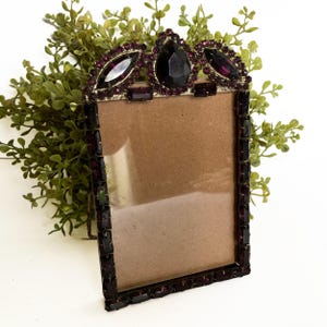 May include: A vintage rectangular photo frame with a decorative crown-like top. The frame is embellished with dark purple gemstones and a gold-coloured metal trim. The frame is empty, ready to display a photo. Green foliage is in the background.