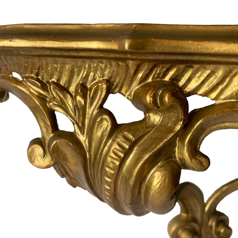 May include: A gold-painted, ornate, decorative wall shelf with intricate scrollwork and floral details. The shelf is made of wood and has a rectangular shape.
