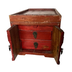 May include: Antique red wooden cabinet with two drawers and a hinged door on each side. The cabinet has a distressed finish, with visible wear and tear. The drawers have metal pulls. The top of the cabinet is a darker brown color.