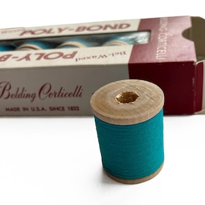 Könnte beinhalten: Eine Holzspule mit türkisfarbenem Faden steht vor einer teilweise sichtbaren Schachtel. Auf der Schachtel steht "POLY-BOND Bel-Waxed" und "Belding Corticelli, Made in U.S.A. Since 1832". Der Faden ist um einen Holzkern gewickelt.