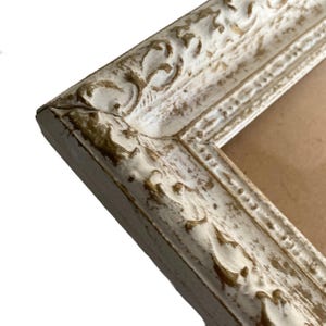 May include: Close-up of a corner of a distressed white picture frame with ornate detailing. The frame has a gold-colored trim and a textured, aged appearance. The frame is angled, revealing a glimpse of the brown backing.