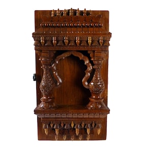 May include: A dark brown wooden wall-mounted shrine with intricate carvings and a small alcove. The shrine has a decorative top with a series of small, pointed finials.