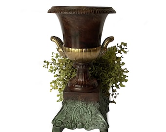 May include: A brown and gold decorative urn with a green base. The urn has a classic design with a round body and a flared top. The base is ornate with a floral pattern.