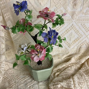 May include: A glass flower arrangement in a green ceramic pot. The flowers are pink, blue, and white and are arranged in a tree shape.