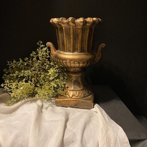 Op de afbeelding: Een goudkleurige decoratieve urn met een gegroefd ontwerp en sierlijke handvatten. De urn staat op een vierkante basis en is geplaatst op een witte doek. Groen is zichtbaar links van de urn. De achtergrond is donker.