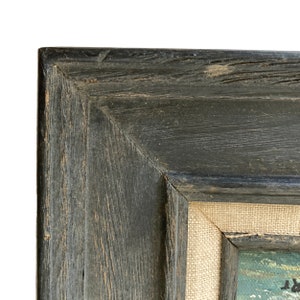 May include: Close-up of a corner of a framed painting. The frame is made of dark, weathered wood with visible grain and texture. The canvas is visible, with a light-colored, woven texture. The painting itself is partially visible, with hints of color.