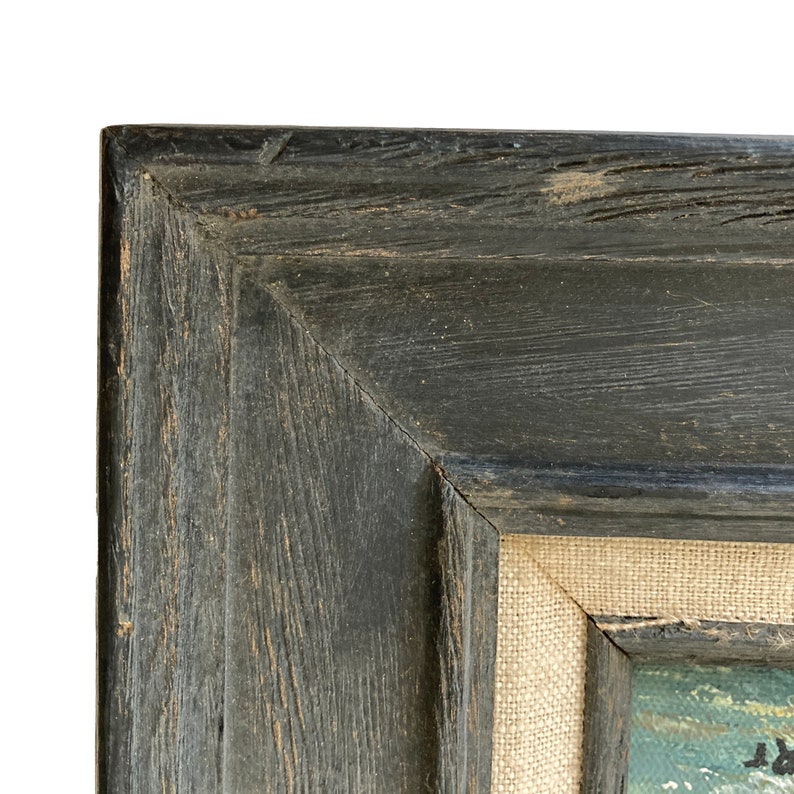 May include: Close-up of a corner of a framed painting. The frame is made of dark, weathered wood with visible grain and texture. The canvas is visible, with a light-colored, woven texture. The painting itself is partially visible, with hints of color.