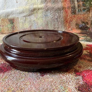 May include: A dark brown wooden pedestal with a smooth, polished finish. The pedestal has a circular top with a small hole in the center and a layered base.