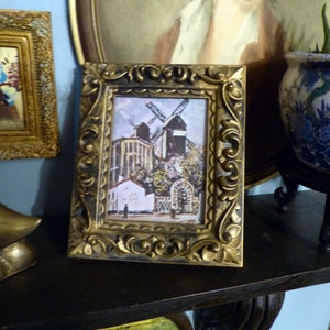 May include: A framed print of a painting depicting a windmill in a village setting. The frame is ornate and gold in color. The print is in a vintage style.
