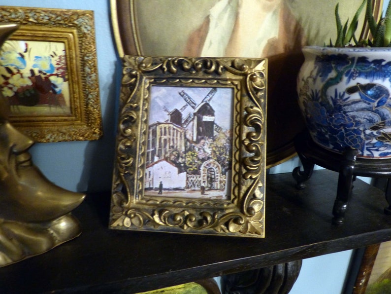 May include: A framed print of a painting depicting a windmill in a village setting. The frame is ornate and gold in color. The print is in a vintage style.