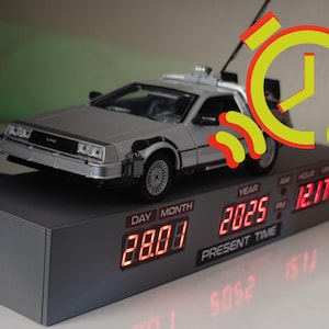 Könnte beinhalten: Ein silbernes DeLorean-Zeitmaschinenmodell sitzt auf einem schwarzen Uhrensockel mit einem roten digitalen Display, das das Datum "28.07" und das Jahr "2025" anzeigt. Die Uhr zeigt auch die Zeit "12:17" an.