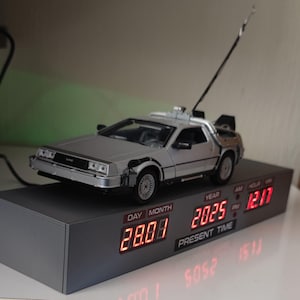 Op de afbeelding: Een zilveren DeLorean tijdmachine replica met een rood digitaal display dat de datum "28.08" en het jaar "2025" laat zien. Het display toont ook de tijd "12:17" met AM/PM-indicatoren.