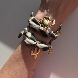 Pode incluir: Pulseiras com pingentes dourados com faixas de tecido estampadas em preto e branco. Uma pulseira apresenta um pingente de caranguejo, enquanto a outra tem um cacto e uma estrela do mar. As pulseiras são usadas no pulso.