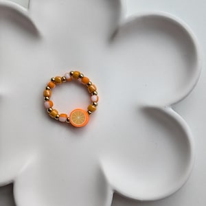 Könnte beinhalten: Ein Perlenring mit einem Orangenscheiben-Design. Der Ring besteht aus kleinen orangefarbenen, gelben, rosa- und goldfarbenen Perlen. Der Ring liegt auf einem weißen, blütenförmigen Teller.