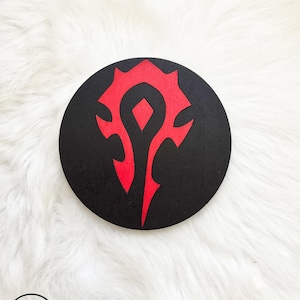 May include: A black and red wooden coaster featuring a stylised symbol of a red eye with a diamond in the centre and a jagged, flame-like design around it.