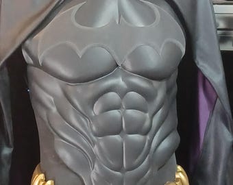 May include: A black costume torso piece featuring a molded muscular chest with a prominent bat-shaped emblem. The garment includes a black cape with a purple inner lining and a metallic gold utility belt with a central circular buckle design.