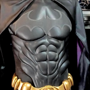May include: A black and gold Batman costume with a sculpted muscle chest and a bat symbol on the chest. The costume includes a black cape and a gold belt with a bat symbol buckle.