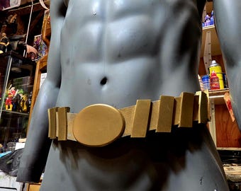 May include: A gold-coloured utility belt with a large central buckle and rectangular compartments. The belt is worn by a grey mannequin. The background includes shelves with various figurines and spray paint cans.