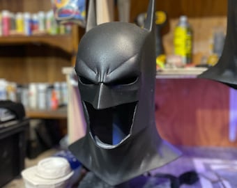 May include: A black Batman mask with a detailed sculpt of the character's face. The mask is sitting on a black surface.