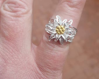 May include: A silver filigree ring with a white flower design and a gold center. The flower is a stylized edelweiss.