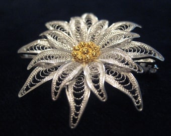 Könnte beinhalten: Eine Brosche aus silbernem Filigran in Form einer Blume mit goldener Mitte. Die Blume hat viele zarte Blütenblätter und ein detailliertes Design.