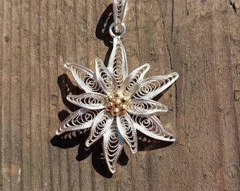 May include: A silver filigree pendant in the shape of an Edelweiss flower with a gold center. The pendant is hanging from a wooden surface.