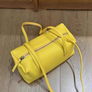May include: A bright yellow handbag with a rectangular shape and a zipper closure. The bag has two handles and a long strap. The bag is made of a textured material.