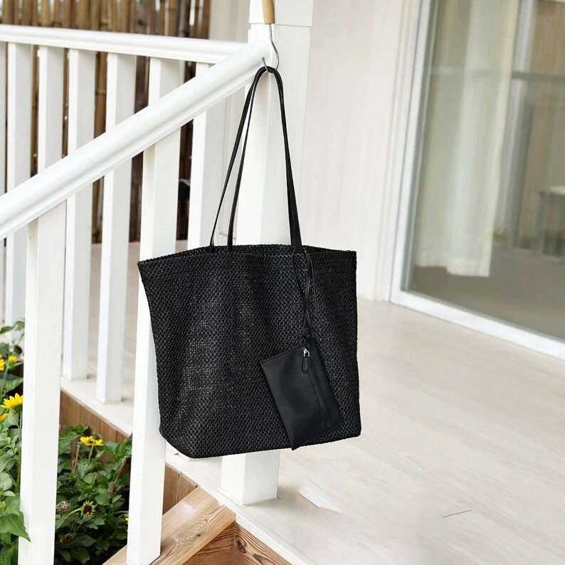 May include: A black woven tote bag with long handles and a small attached pouch. The bag is hanging from a hook on a white railing. The bag has a rectangular shape and a textured surface.