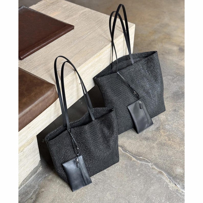 May include: Two black woven tote bags with long, thin handles. Each bag has a small, matching pouch attached. The bags are made of a textured material and are displayed on a neutral surface.
