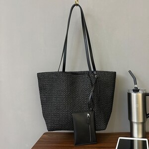 May include: A black woven tote bag with long black handles and a small matching pouch. The bag is rectangular and has a textured appearance. The pouch has a zipper closure and a strap. The bag is hanging from a gold hook.