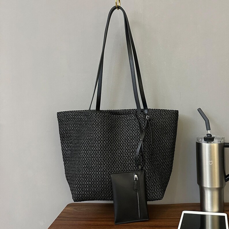 May include: A black woven tote bag with long black handles and a small matching pouch. The bag is rectangular and has a textured appearance. The pouch has a zipper closure and a strap. The bag is hanging from a gold hook.