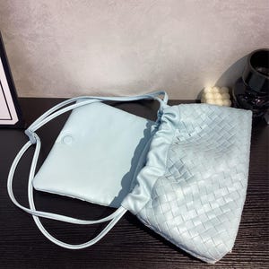 May include: A light blue leather crossbody bag and a matching pouch. The crossbody bag features a woven design on the main body and a drawstring closure. The pouch is rectangular with a button closure and a long strap.