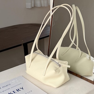 May include: Cream-colored rectangular handbag with long, thin straps. The bag has a silver zipper closure and a matching zipper pull. The bag is sitting on a white surface, with a mirror reflecting the bag's image.
