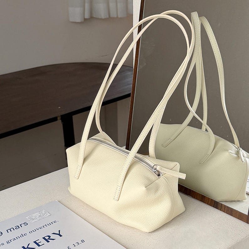 May include: Cream-colored rectangular handbag with long, thin straps. The bag has a silver zipper closure and a matching zipper pull. The bag is sitting on a white surface, with a mirror reflecting the bag's image.