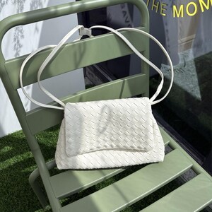 May include: A white woven crossbody bag with a long strap rests on a green chair. The bag features a rectangular shape with a flap closure. The chair is a light green color and is positioned on a patch of green grass.