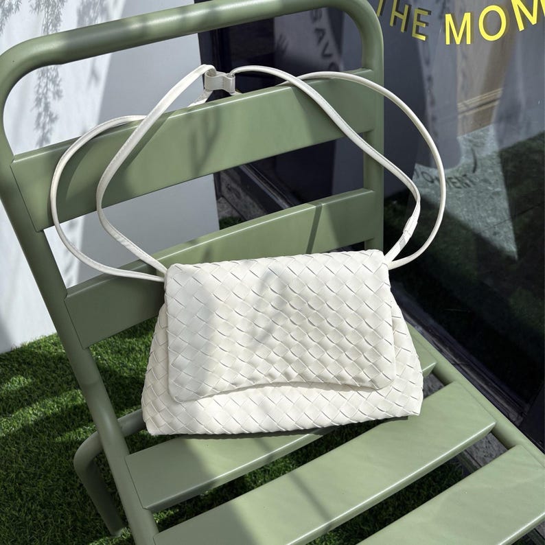 May include: A white woven crossbody bag with a long strap rests on a green chair. The bag features a rectangular shape with a flap closure. The chair is a light green color and is positioned on a patch of green grass.