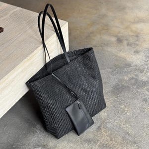 May include: A black woven tote bag with long, thin handles. A smaller, matching black pouch with a zipper is attached. The bag and pouch are made of a textured material, and the background is a neutral color.