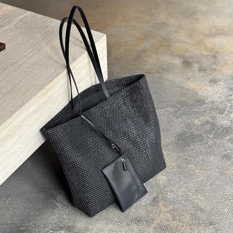 May include: A black woven tote bag with long, thin handles. A smaller, matching black pouch with a zipper is attached. The bag and pouch are made of a textured material, and the background is a neutral color.