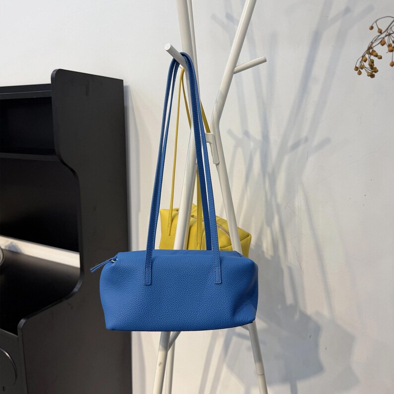 May include: A vibrant blue rectangular handbag with long, thin straps. The bag is made of textured material and has a zipper closure. A yellow bag hangs in the background. The bags are hanging on a white coat rack.