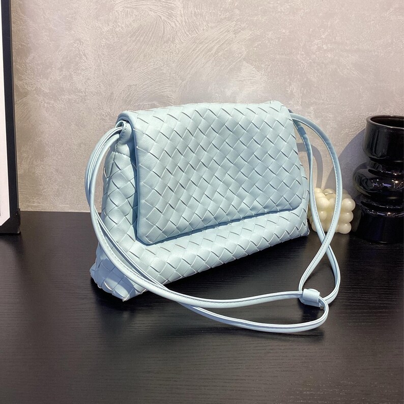 May include: A light blue crossbody bag with a woven design. The bag features a flap closure and a long, thin strap. The bag is made of a soft material and is designed to be worn over the shoulder.