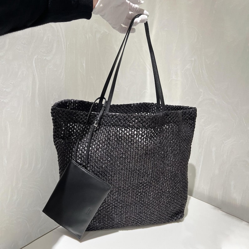 May include: Black woven tote bag with long shoulder straps and a small matching pouch. The bag has a rectangular shape and a textured, open weave design. The pouch is smooth and rectangular.