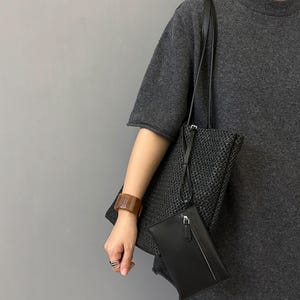 May include: A black woven tote bag with a long shoulder strap, paired with a black leather clutch. A brown rectangular bracelet is worn on the wrist. The items are set against a gray background.
