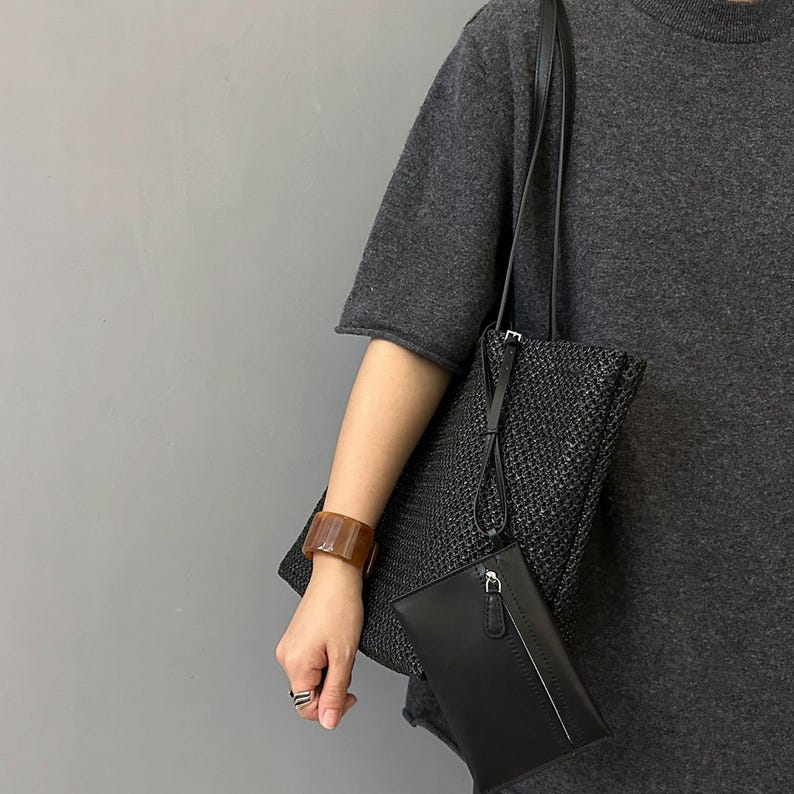 May include: A black woven tote bag with a long shoulder strap, paired with a black leather clutch. A brown rectangular bracelet is worn on the wrist. The items are set against a gray background.