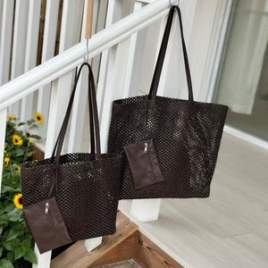 May include: Two brown woven tote bags with matching small pouches hanging on a white railing. The bags have long, dark brown straps. The bags are made of a woven material with a grid-like pattern. The pouches have a zipper closure.