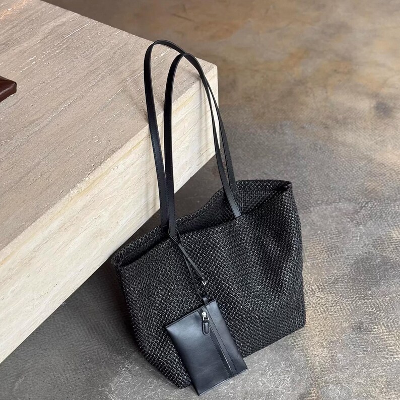 May include: Black woven tote bag with long black handles and a small matching pouch. The bag is made of a textured material and has a rectangular shape. The pouch is attached to the bag with a thin strap.