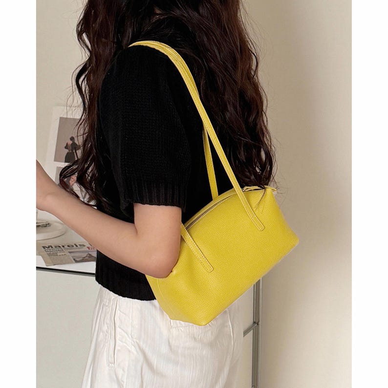 May include: A bright yellow rectangular handbag with a zipper closure and long straps. The bag is made of a textured material and is being carried over the shoulder. The bag is a vibrant accessory.