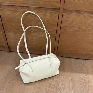 May include: A cream-colored handbag with a rectangular shape and a zipper closure. The bag has two long, thin handles and is made of a textured material. The bag is sitting on a wooden floor.
